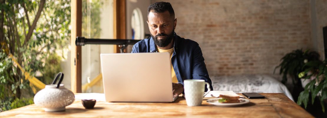 Man with moustache and beard wearing navy blue button-down shirt over a yellow t-shirt using laptop computer at a wooden table in his home.