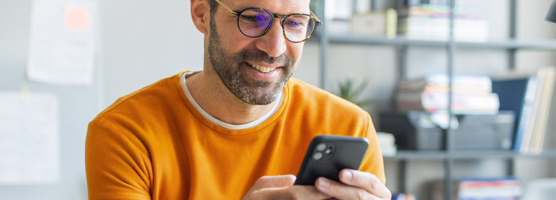 Lightly bearded man wearing tortoiseshell glasses and orange sweatshirt smiling as he holds up and looks at his cellphone.