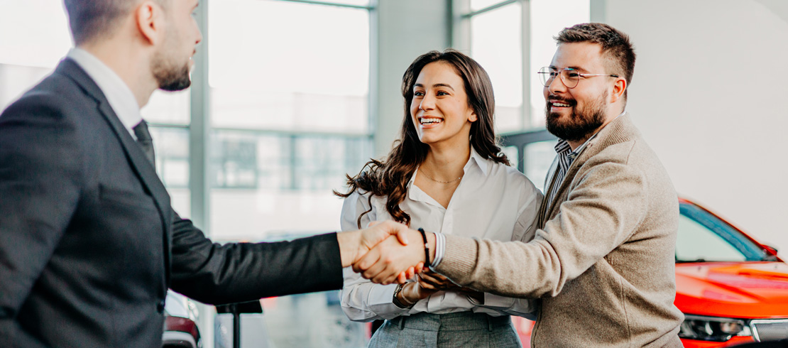 man shaking salesperson's hand with his female partner at a car dealership